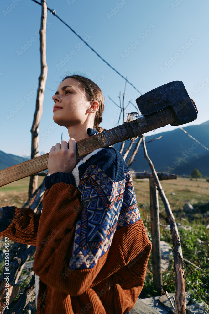 Fototapeta premium woman sledgehammer mallet rural vineyard mountains farmer outdoor carrying heavy wooden tool on shoulder, confident young agricultural worker in patterned jacket among trellis and fields under blue