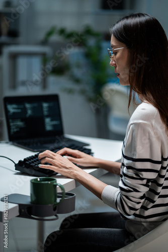 Focused Asian programmer wearing glasses, typing on a keyboard at late night in the office. Software development, cybersecurity, remote work, web design.  Freelance working overtime.