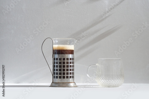 French press filled with freshly brewed coffee standing beside an empty textured glass mug on a bright surface with soft natural light and long shadows against a neutral background. Sunny day