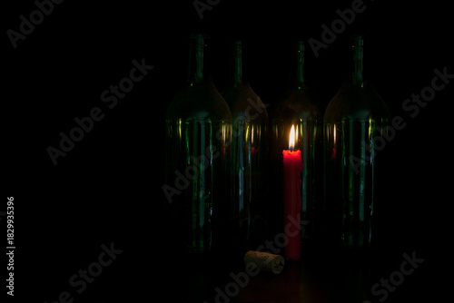 Against a deep black backdrop, dusty wine bottles stand in the warm glow of a red candle. Its light gently reflects in the glass, lending the still life a quiet, poetic dignity.