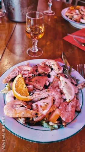 Close-up view of a gourmet seafood salad featuring large prawns, tomato slices, and fresh greens on a white plate. A glass of amber alcohol rests on the wooden dining table in the background.