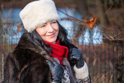 Woman in fur coat and hat smiles warmly outdoors in winter sunlight, enjoying the season while surrounded by nature, embodying a cozy and cheerful atmosphere