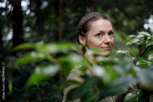 A woman with brown eyes and braided hair smiles softly, framed by lush green leaves in a sun-dappled forest. Soft, natural light filters through the canopy, creating a serene, earthy atmosphere.