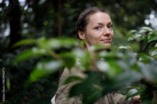 A woman with brown eyes and braided hair smiles softly, framed by vibrant green leaves in a lush, dimly lit forest. She wears a beige quilted jacket, with soft natural light filtering through the cano