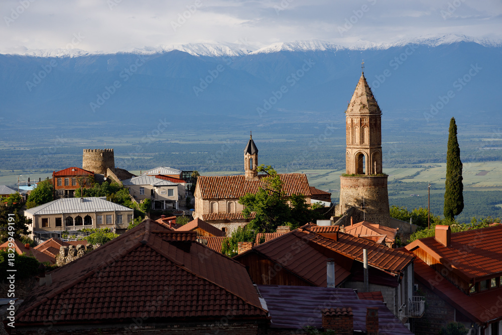 Fototapeta premium Sunlit village with terracotta roofs and stone towers, nestled against snow-capped mountains under a partly cloudy sky.