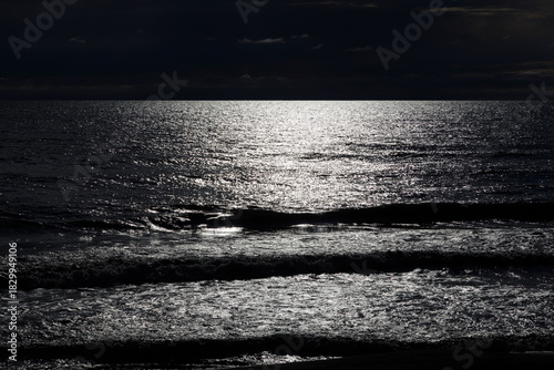 Dark ocean under a stormy sky, with a brilliant, shimmering light path reflecting across the water's surface, creating a stark contrast against deep shadows.