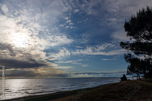 Dramatic sky with white clouds and sunbeams over a calm sea. A lone figure in a red hat sits on a grassy shore beside dark pine trees, silhouetted against the vast horizon.