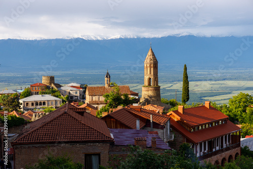 Sunlit village with terracotta roofs and stone towers, nestled against snow-capped mountains under a soft, cloudy sky. Lush greenery and expansive valleys stretch below.