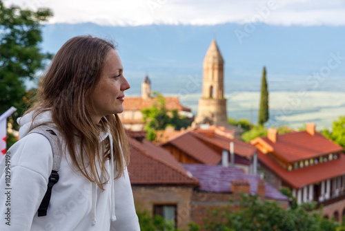A woman in a white hoodie gazes at a scenic town with red-tiled roofs and a stone tower, set against distant mountains under soft, diffused daylight.