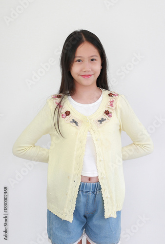 Happy young Asian girl standing against white background, casually posing in pastel yellow cardigan smiling sweetly at camera.