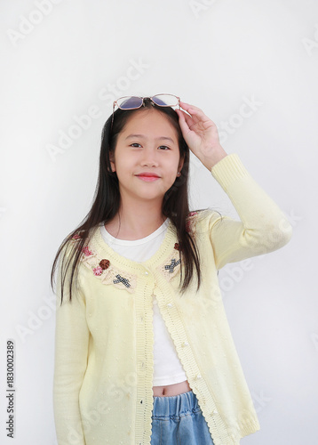 Trendy young Asian girl in pastel yellow cardigan against white background, pushing her stylish eyeglasses up onto her head while smiling at camera.