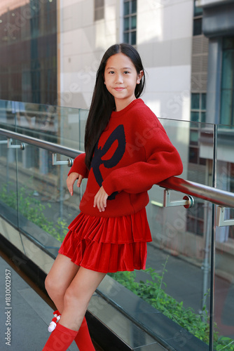 Portrait of happy young Asian girl with long hair, wearing festive red sweater and skirt, smiling and leaning against glass railing in modern city background.