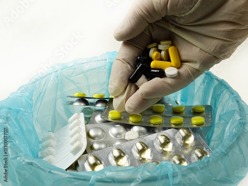 A hand in a white rubber glove pours expired pills into a container with expired pill blister packs medicines
