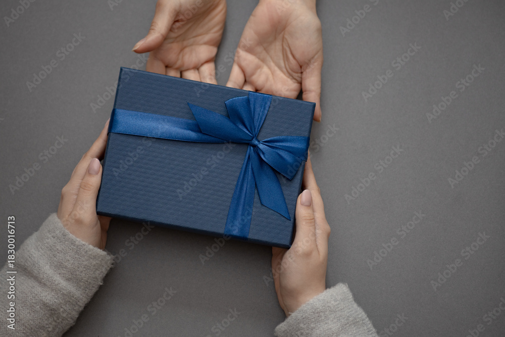 Naklejka premium Top view of male and female hands holding blue gift box with ribbon on gray background Flat lay. Present for birthday, valentine day, Christmas, New Year, copy space