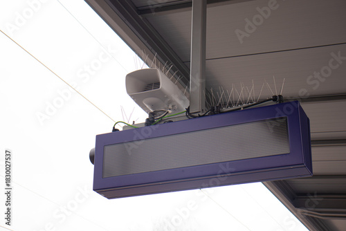Blank digital display board with bird spikes mounted under a station roof