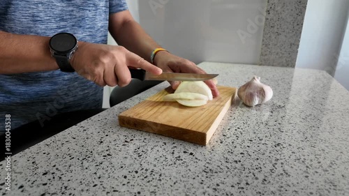 A person's hands chop an onion with a knife on a wooden board to prepare food