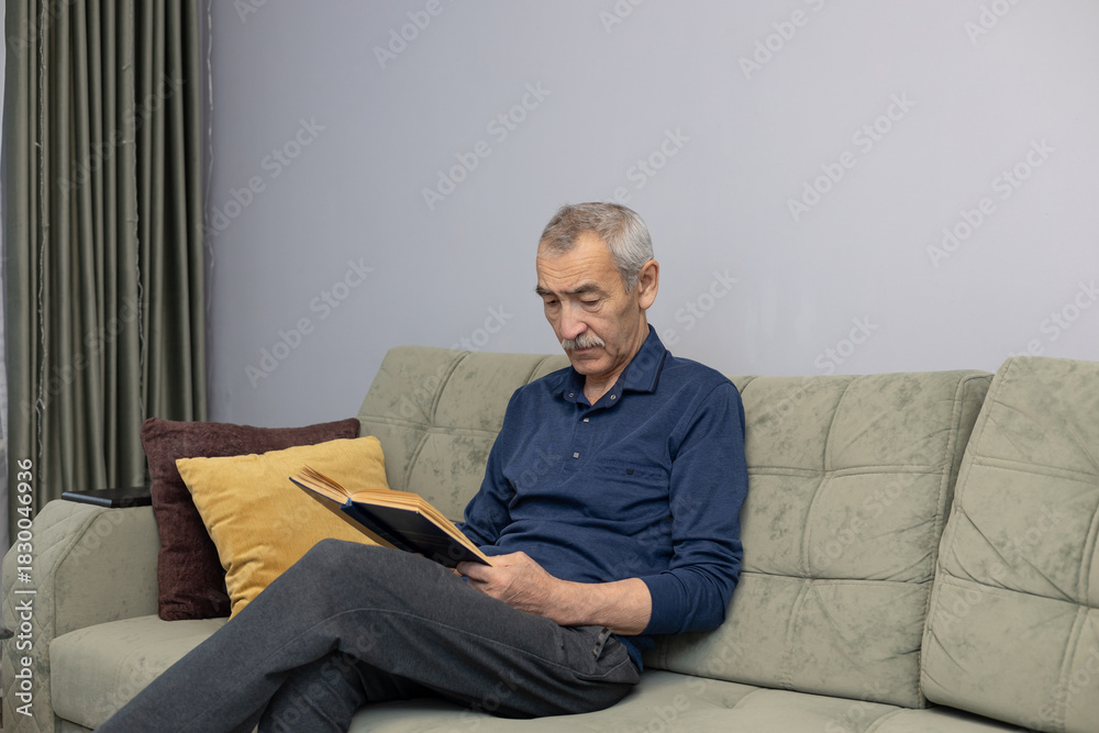 Fototapeta premium An elderly Asian man reading a book while sitting on the sofa at home