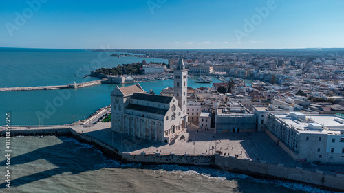 A panoramic coastal view of Trani shows the famous seaside cathedral overlooking turquoise waters, a long breakwater with lighthouses, and the historic old town bathed in bright Mediterranean sunlight
