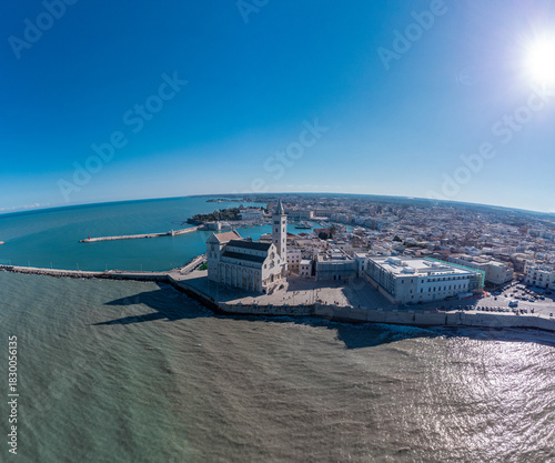 A panoramic coastal view of Trani shows the famous seaside cathedral overlooking turquoise waters, a long breakwater with lighthouses, and the historic old town bathed in bright Mediterranean sunlight