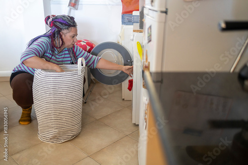 Plus size woman with dreadlocks doing laundry, reaching into a washing machine door next to a large laundry basket.