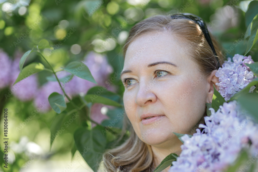 Fototapeta premium A blonde woman stands near blooming lilacs in a city park. Summer.