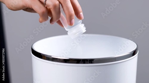 Close Up Of Hand Disposing Small White Pills From Clear Plastic Container Into White Trash Can With Silver Rim On Gray Background Studio Lighting