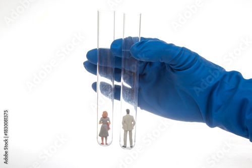 A gloved hand holds two test tubes containing miniature figurines of a man and woman, symbolizing human experimentation or scientific research on a white background.