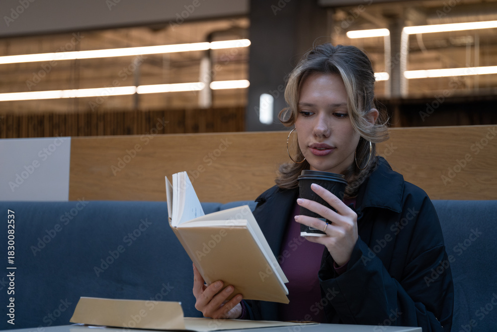 Fototapeta premium Young woman reads a book while holding a takeaway coffee cup in a cafe setting