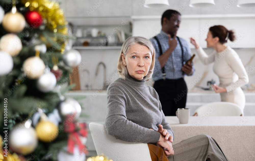Obraz premium Upset old-aged woman sitting in the kitchen next to Christmas-tree and middle-aged man and woman arguing standing behind