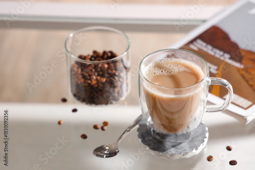 Glass cup of cappuccino with coaster and coffee beans on table in room, closeup