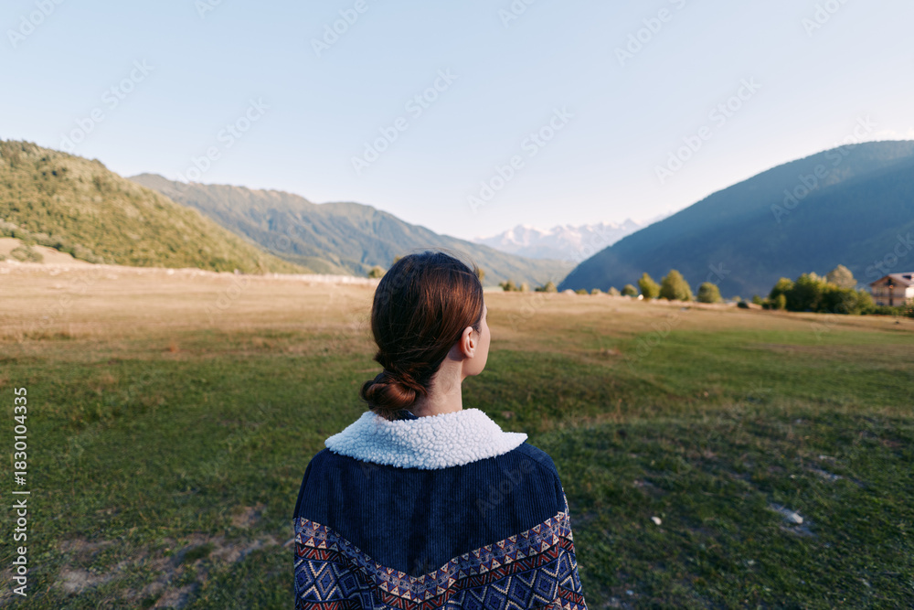Fototapeta premium Woman back in sweater stands in wide valley landscape facing distant mountains, nature travel scene with serene meadow and alpine ridges, outdoor exploration and peaceful solitude.