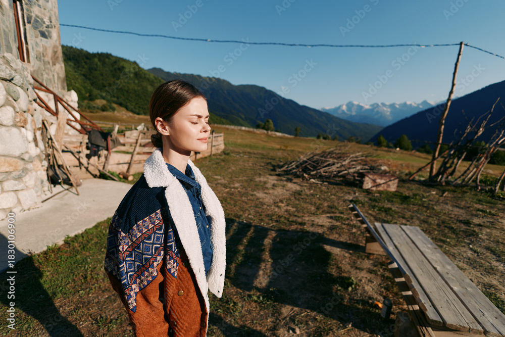 Naklejka premium Woman portrait in sunlight by rustic cabin with closed eyes, standing in nature overlooking mountains and meadow. Serene outdoor scene with jacket, calm expression and peaceful landscape.