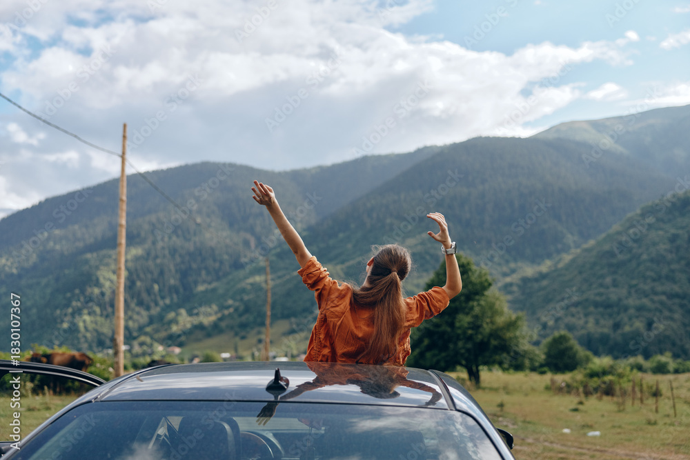 Fototapeta premium Woman stands on a car roof with arms raised enjoying freedom during a road trip in a scenic mountain landscape outdoors, conveying adventure and happiness on the open road