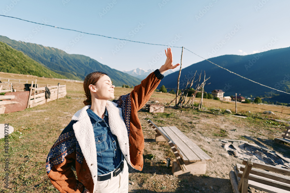 Naklejka premium Woman in denim and warm jacket smiling outdoors in countryside, shielding eyes from sunlight while standing in a meadow with mountains in the background, travel and nature joy.