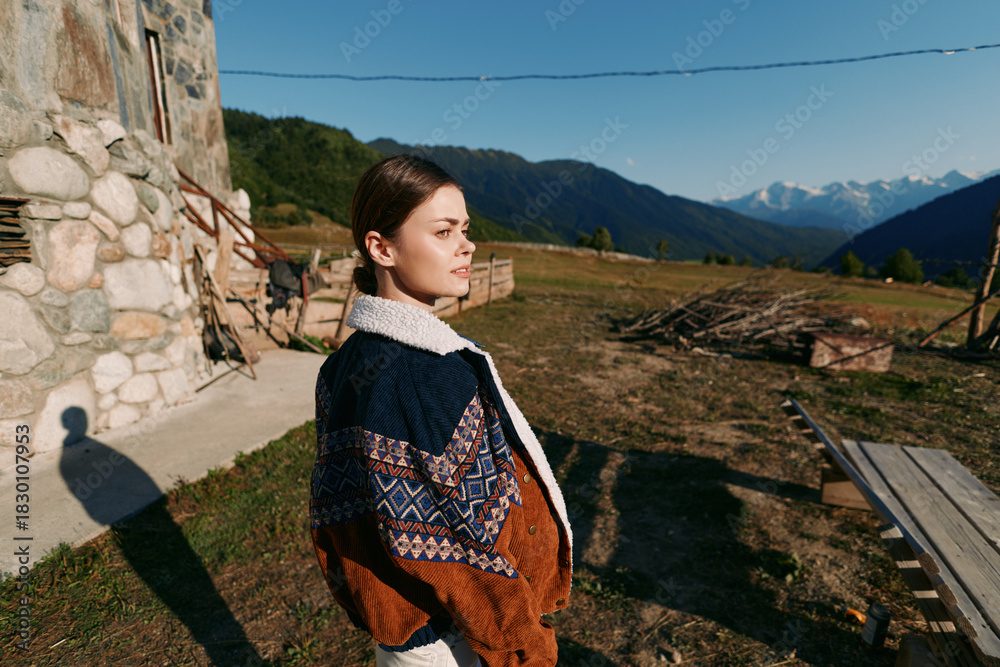 Naklejka premium Woman portrait in countryside wearing patterned sweater near stone cottage, looking toward distant mountains at golden hour. Outdoor lifestyle, travel, nature and peaceful rural scene.