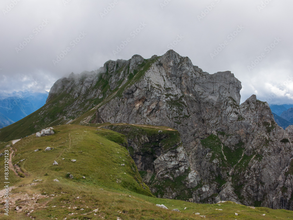Obraz premium A panoramic alpine landscape of Montgart in the Alps featuring rugged rocky cliffs, soft green slopes, a winding mountain road and a dramatic cloudy sky