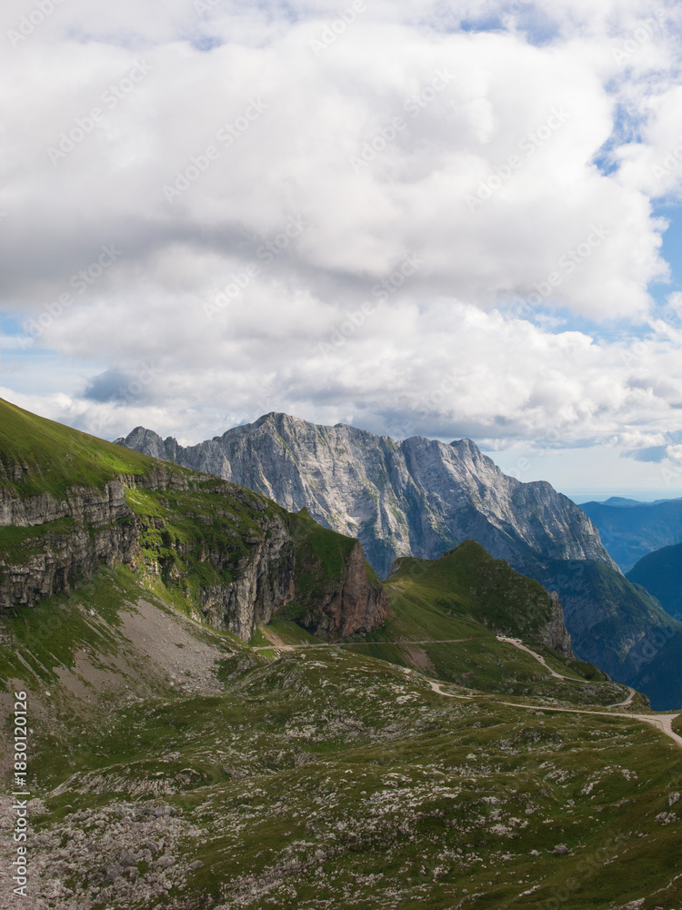 Fototapeta premium A panoramic alpine landscape of Montgart in the Alps featuring rugged rocky cliffs, soft green slopes, a winding mountain road and a dramatic cloudy sky
