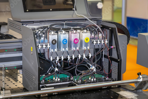 An industrial inkjet printer with the ink system exposed, showing the CMYK cartridges and tubes. The internal mechanism is visible, showing the distribution of ink on the print bed