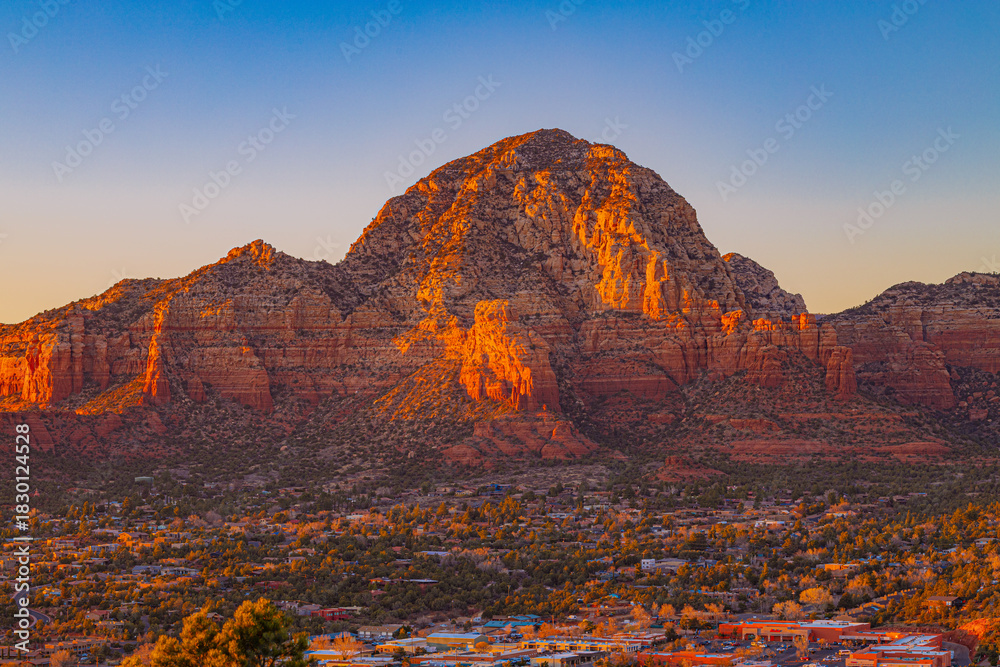 Naklejka premium A warm sunset illuminates the iconic red rock formations of Sedona, Arizona. The glowing cliffs rise above the town below, creating a striking desert mountain landscape.