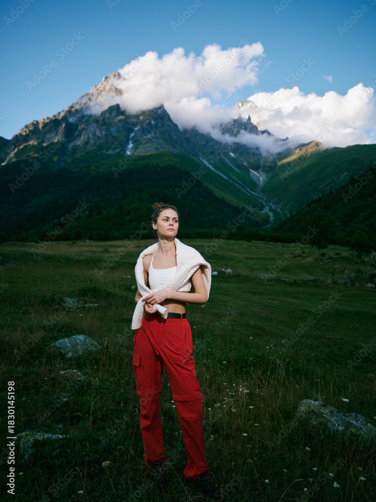 Naklejka premium Woman standing in a grassy meadow with red pants and a draped sweater, alpine mountains and cloudy blue sky behind, capturing outdoor exploration, nature, and expansive landscape