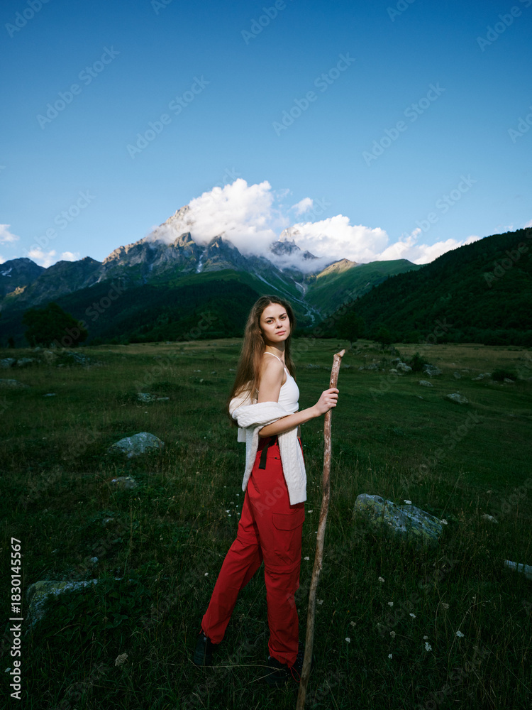 Naklejka premium Woman standing in a green meadow with a wooden staff, red pants and white top, mountains and blue sky behind for an outdoor nature scene