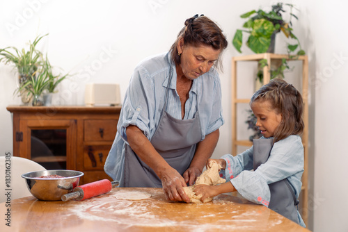 Elderly woman and young girl in aprons kneading dough on table in home kitchen enjoying time together, learning baking skills and sharing joyful family moment