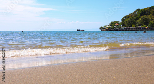Sunny tropical beach view with small waves on golden sand and island resort backdrop