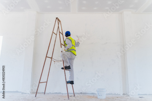 Professional Painter on Ladder Applying Paint to White Wall in Empty Room with Construction Safety Gear and Equipment