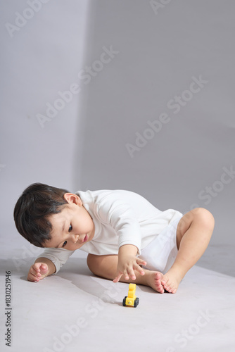 A young child in white clothing lies on the stomach, reaching forward toward colorful toy cars on a plain studio floor.