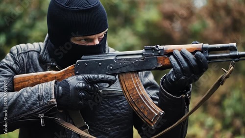 Man wearing black balaclava and leather jacket holding an assault rifle outdoors in nature