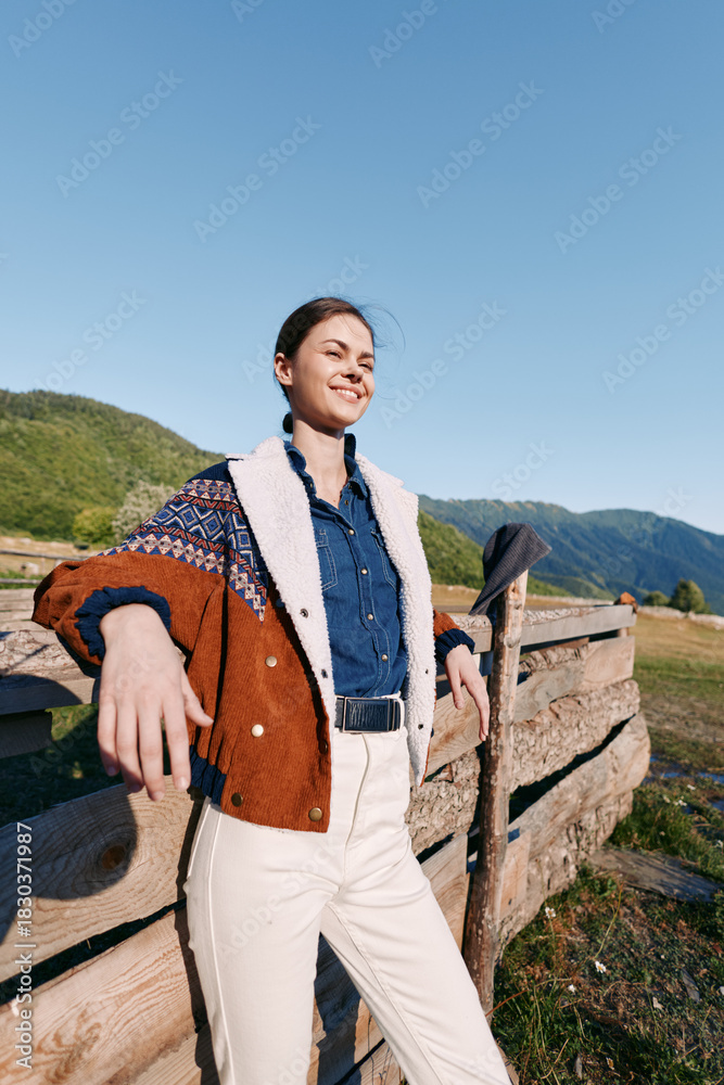 Naklejka premium woman countryside fence mountains jacket smiling outdoors portrait. Young woman stands leaning on a wooden fence in a rural landscape wearing a patterned jacket, smiling confidently under clear blue