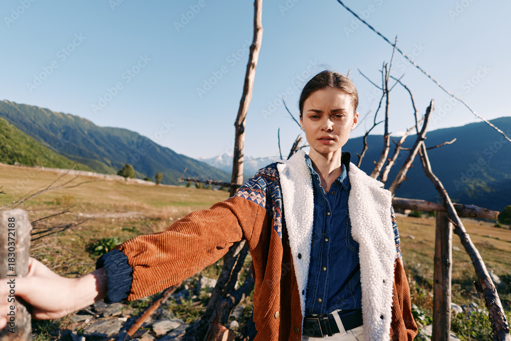 Naklejka premium Woman portrait in mountains countryside by wooden fence wearing warm jacket and denim shirt, standing outdoors with fashion look and scenic valley landscape under clear sky.