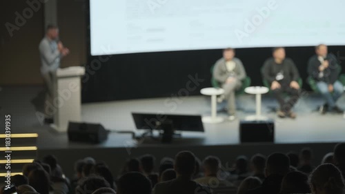 Speaker at podium addresses panel during conference. Blurred audience watches in large theater ambiance. Business discussion.