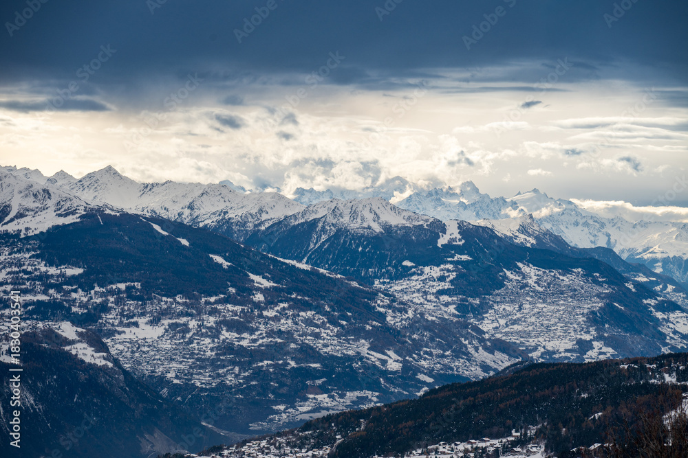 Fototapeta premium Panorama hivernal des Alpes avec chaîne de montagnes enneigées. Ciel nuageux dramatique, ambiance froide et montagneuse.